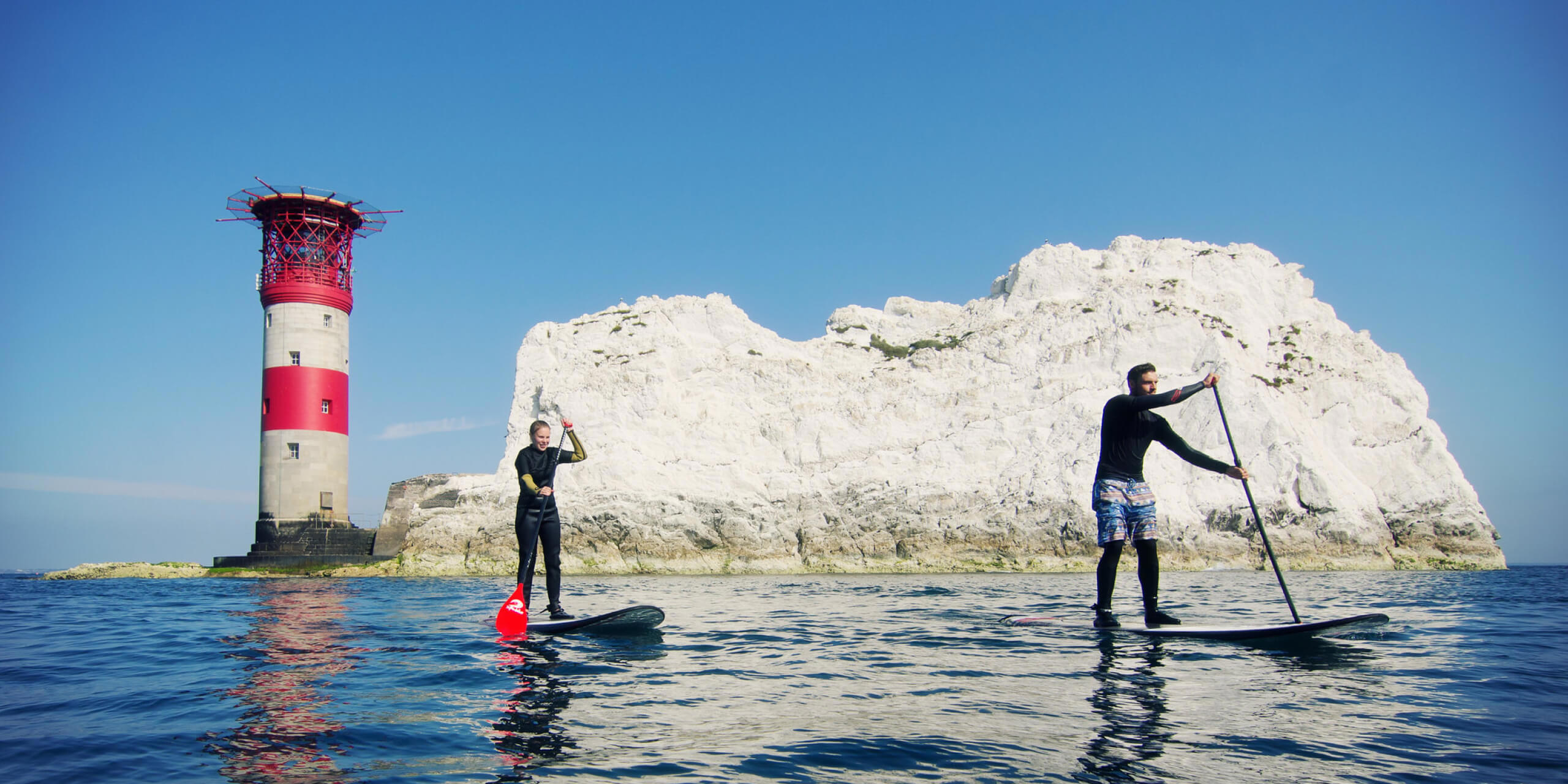 Photo of two people paddle boarding in the ocean in front of a lighthouse